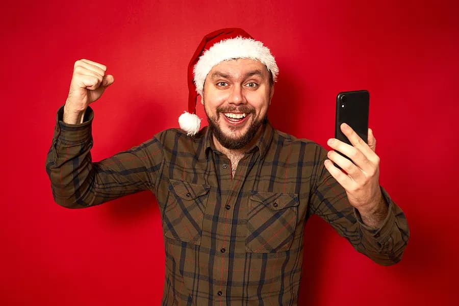 Homem adulto com um chapéu de Papai Noel, animado, segurando um celular