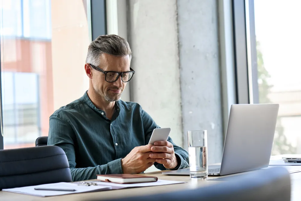 homem olhando para o smartphone enquanto trabalha no laptop