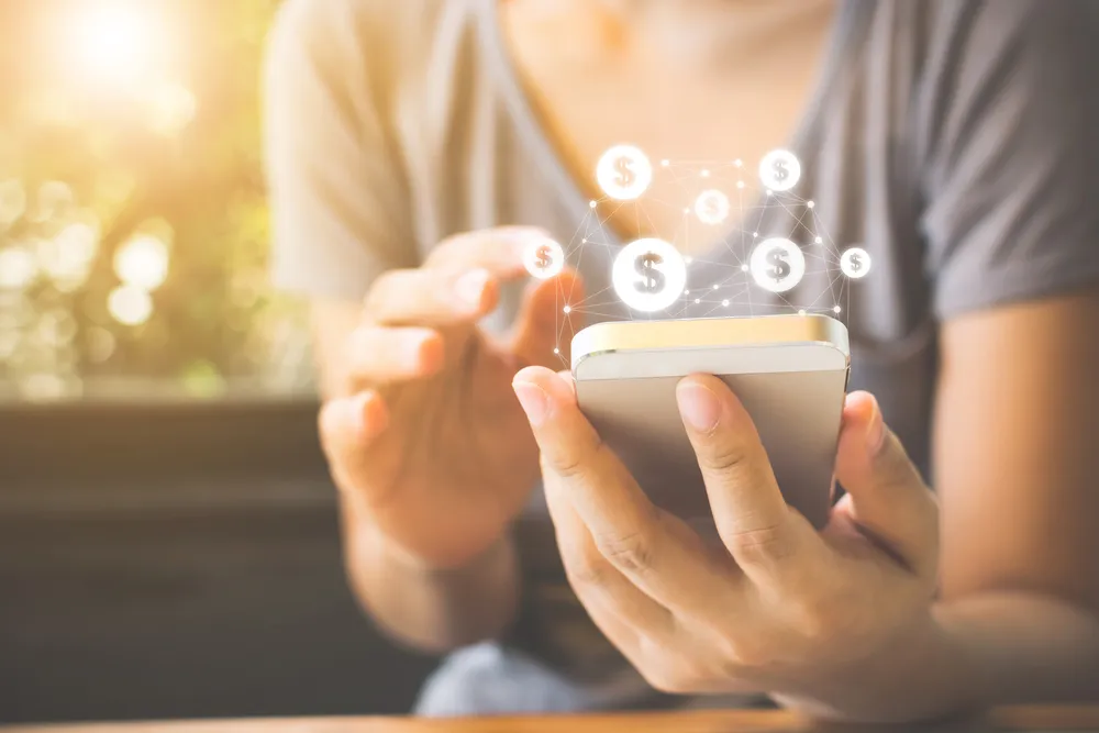 O tronco de uma pessoa segurando um celular, cercado por símbolos de dólar