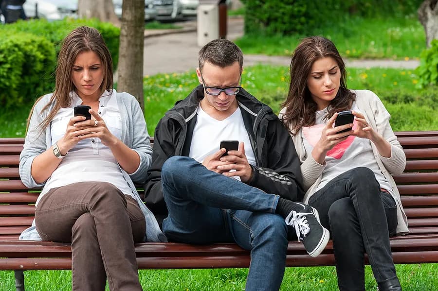 Três pessoas sentadas em um banco de parque, ocupadas com seus celulares.