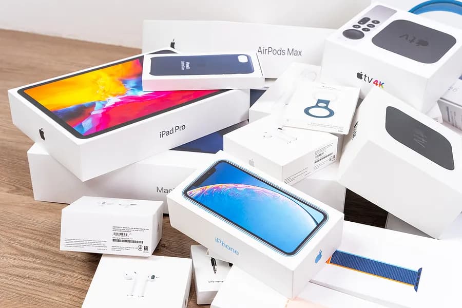 Uma pilha de caixas de produtos da Apple, incluindo iPhone, iPad Pro, AirPods, Apple Watch e Apple TV, dispostas sobre uma superfície de madeira.