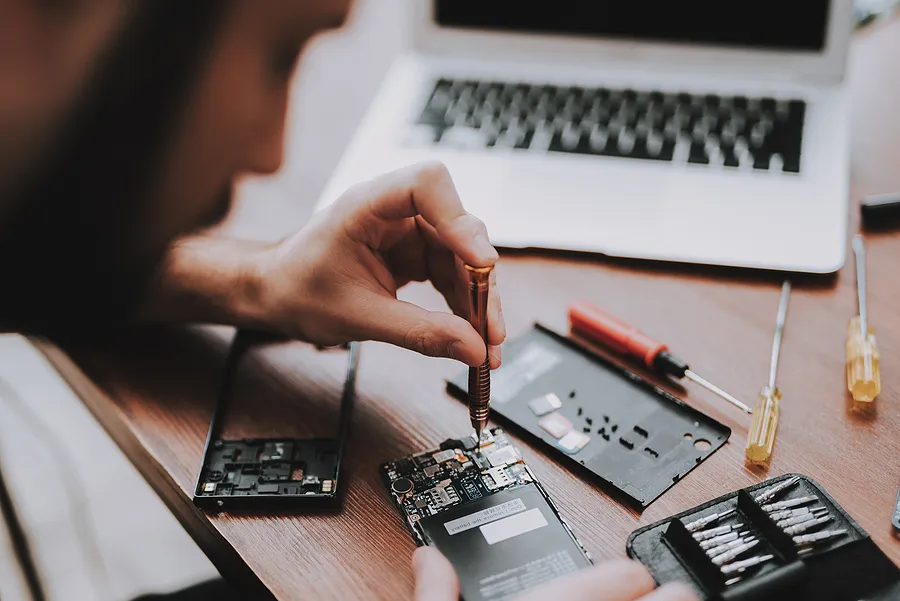 Um técnico usa uma chave de fenda de precisão para consertar os componentes internos de um smartphone em uma mesa de madeira, cercado por pequenas ferramentas, peças de reposição e um laptop aberto.
