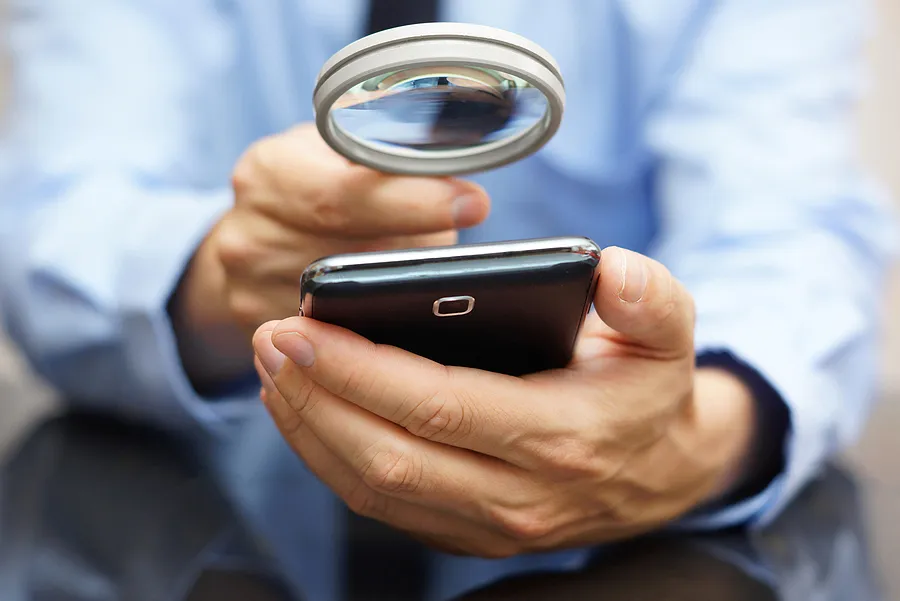 Uma pessoa examinando um smartphone de perto com uma lupa, verificando se o aparelho é original ou se apresenta algum problema.