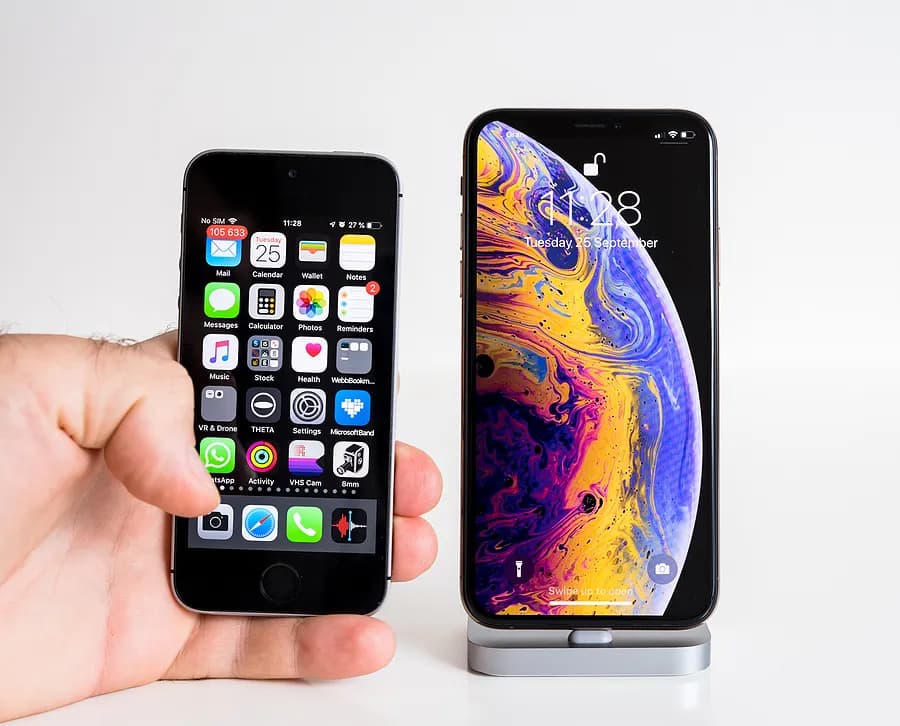 Mão segurando um iPhone antigo e pequeno ao lado de um iPhone moderno e maior em um suporte, mostrando as diferenças de tamanho e design entre um iPhone antigo e um modelo mais recente.